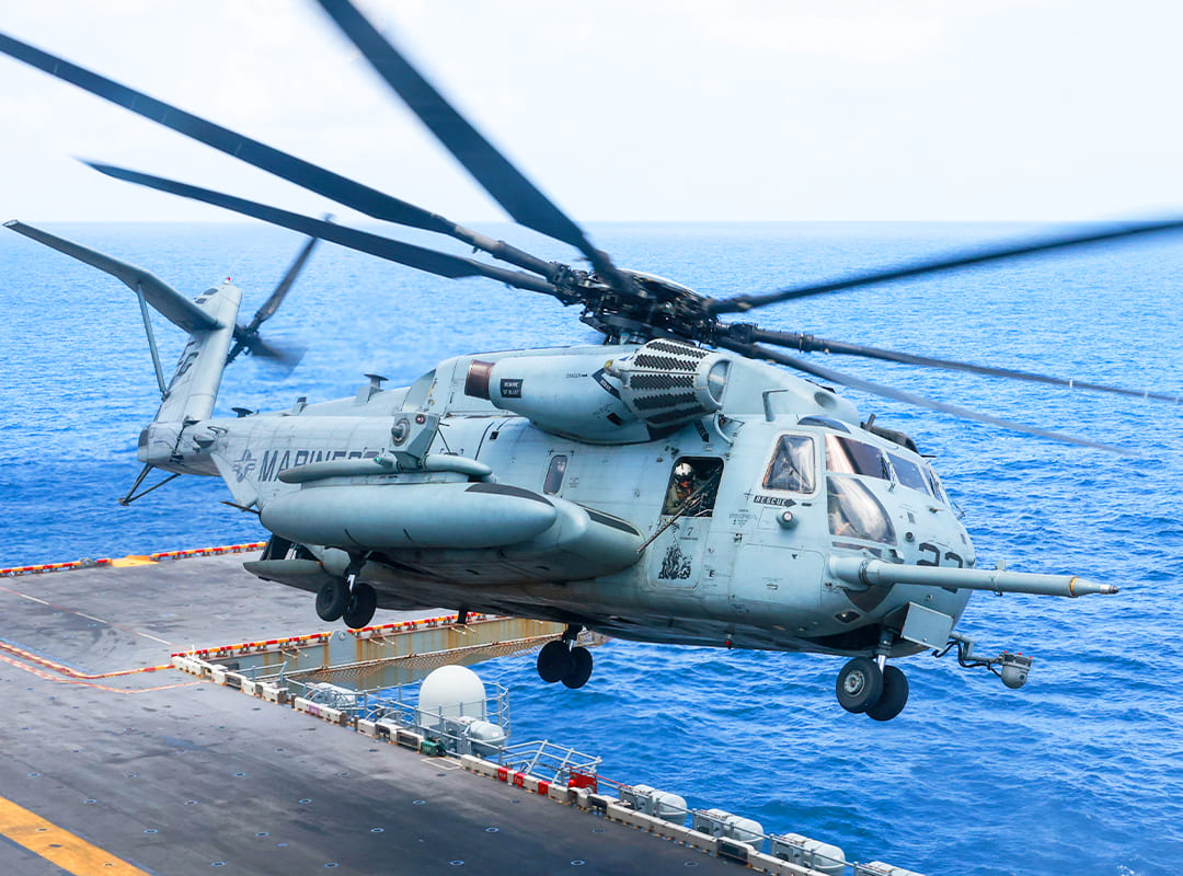 USMC CH-53 Super Stallion helicopter taking off at an angle from an aircraft carrier in the ocean.