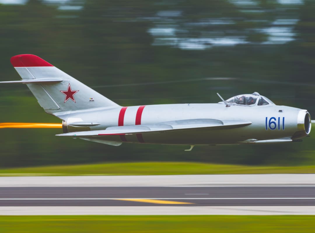 A MiG-17 flies low and fast over a roadway with trees in the background, and a flame shooting from the back engine.