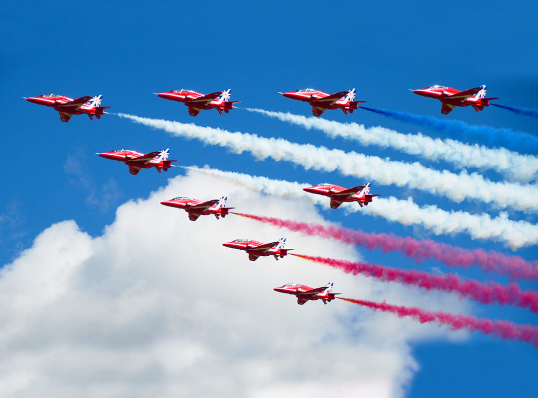 The Royal Air Force Red Arrows demonstration team, with all nine ships flying against a blue sky with clouds, and their red, white and blue smoke trails coming from the tails.