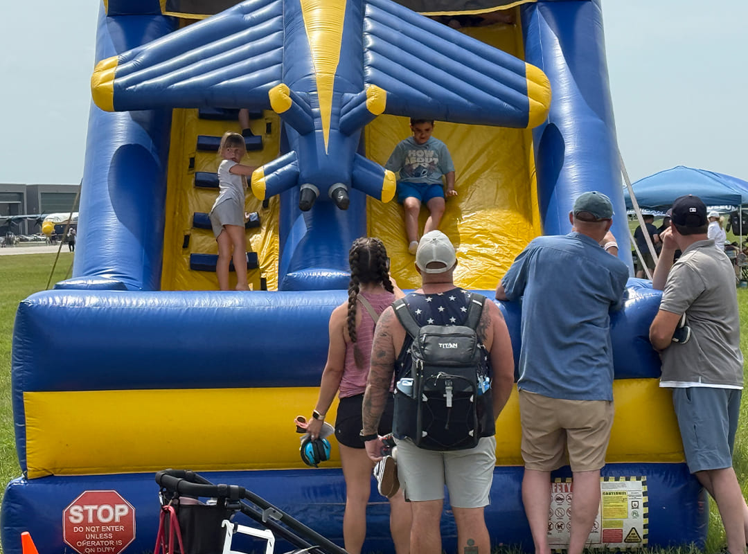 Children climb and slide on a large blue-and-yellow inflatable aircraft-themed bounce house while adults watch from the ground in the Adventure Zone at Thunder Over Michigan.