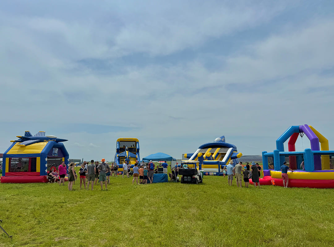 Families gather around multiple colorful inflatable attractions and bounce houses spread across a grassy field in the Adventure Zone at Thunder Over Michigan.