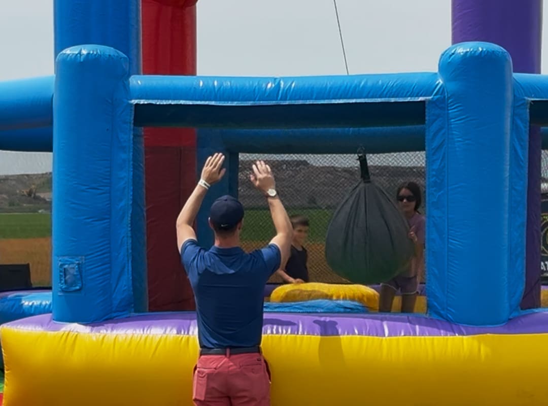 Children play inside a colorful inflatable bounce house with a hanging obstacle while an adult stands outside supervising in the Adventure Zone at Thunder Over Michigan.