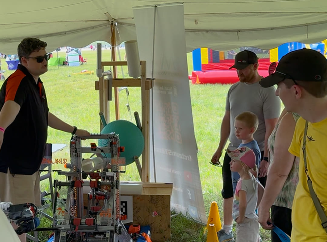 A presenter demonstrates a robotics project under a tent while families and children watch and learn at the Exploration Station during Thunder Over Michigan.