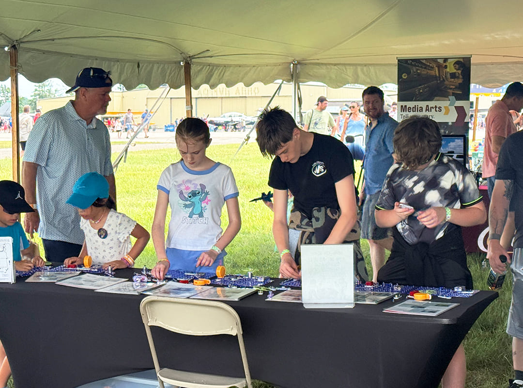 Children and adults participate in a hands-on STEM activity, assembling electronic components and circuits at a table inside the Exploration Station tent.