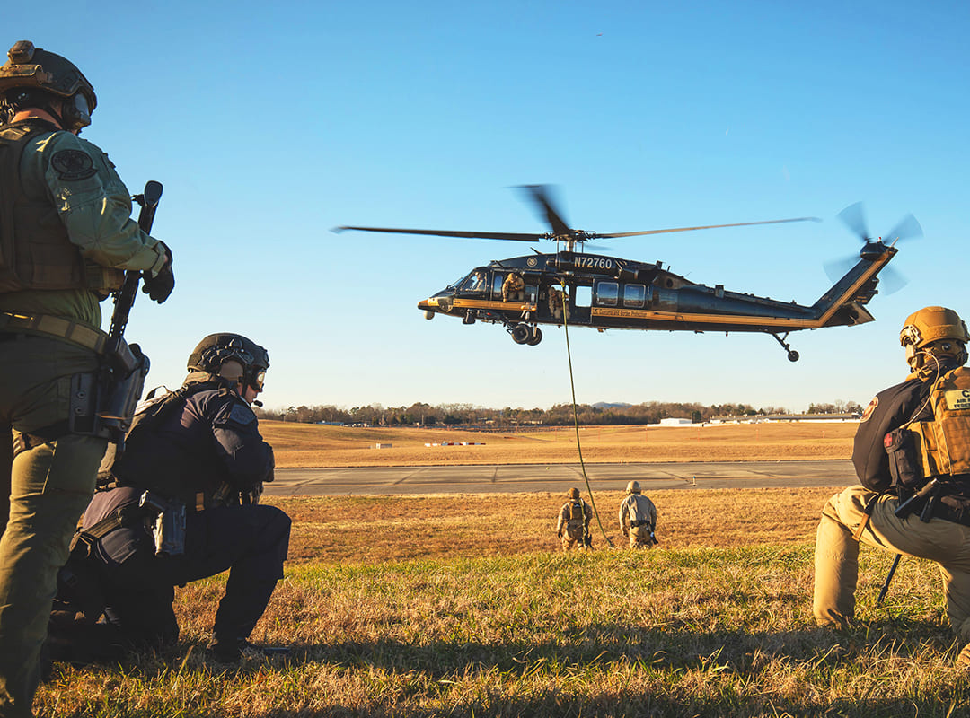 Tactical personnel prepare for a fast-rope training exercise as a Black Hawk helicopter hovers low over an airfield and two team members descend to the ground.