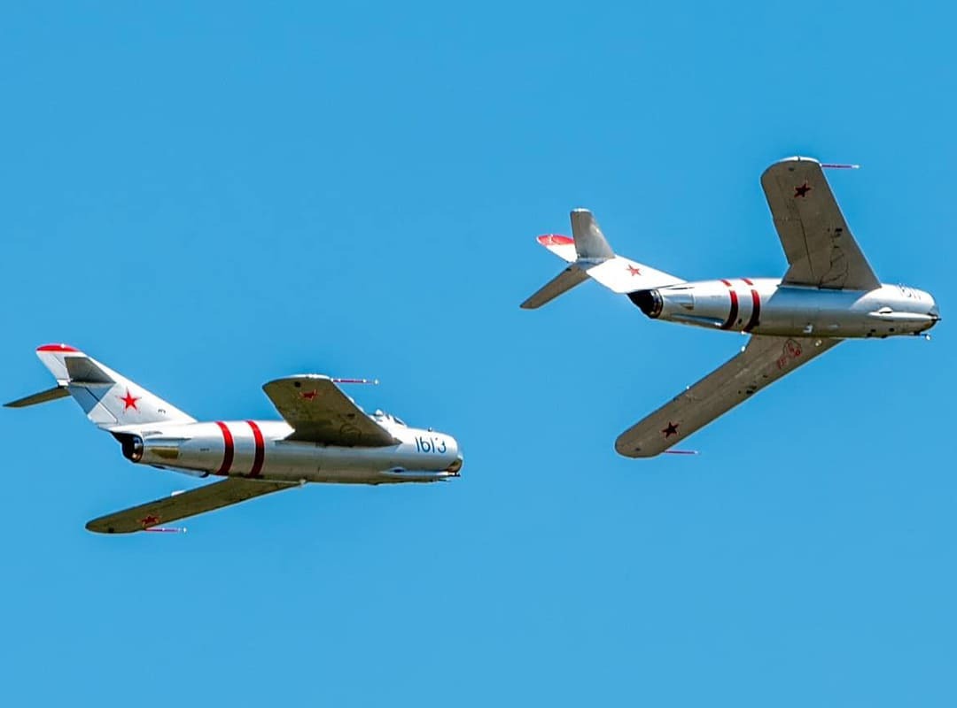 Two silver MiG jet aircraft with red star markings fly in formation against a clear blue sky.