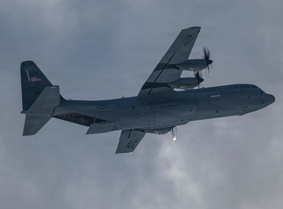 A U.S. Air Force C-130 Hercules transport aircraft flies beneath an overcast sky with all four propellers turning.