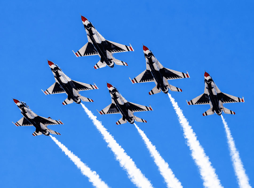 Six U.S. Air Force Thunderbirds jets climb in formation, leaving bright white smoke trails against a clear blue sky.